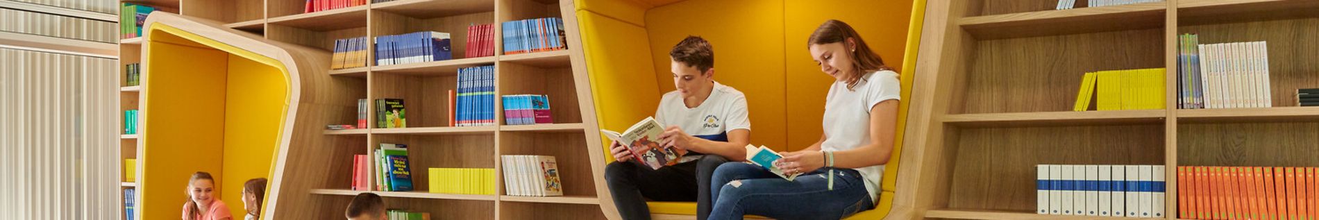 Bibliothek der Stadtteilschule Lurup, Holzbänke und gepolsterte Sitze, Schülerinnen und Schüler lesen und unterhalten sich.